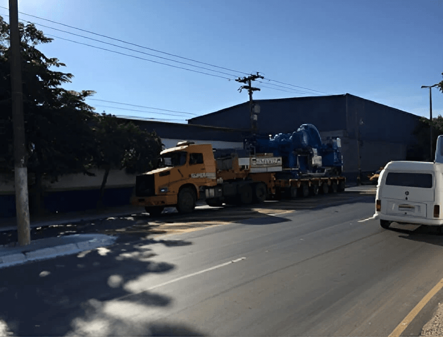 Caminhão amarelo realizando o transporte de grande equipamento industrial azul em frente a galpão metálico.