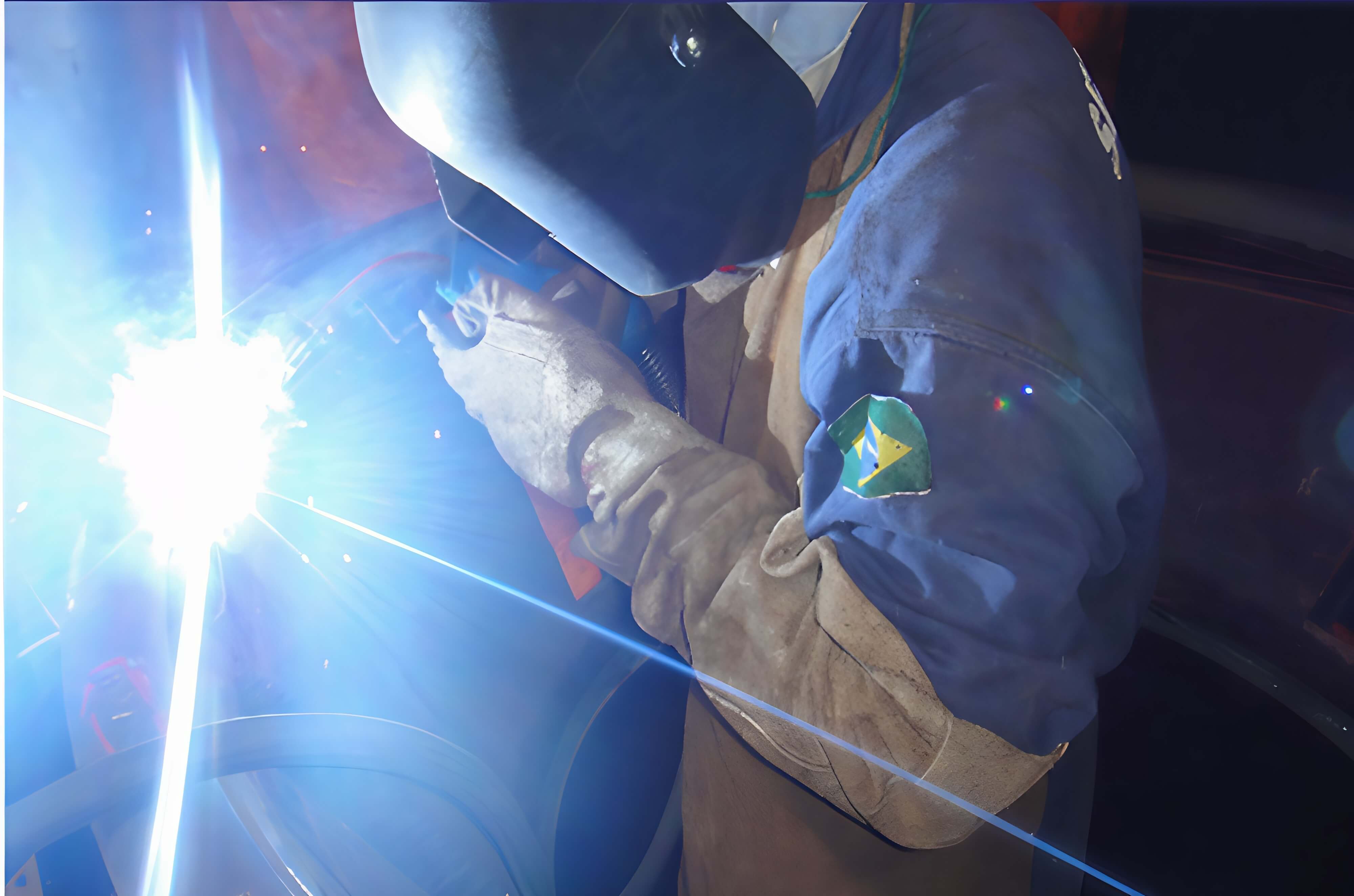 Soldador em ação com máscara de proteção, emitindo faíscas azuis intensas durante processo industrial.