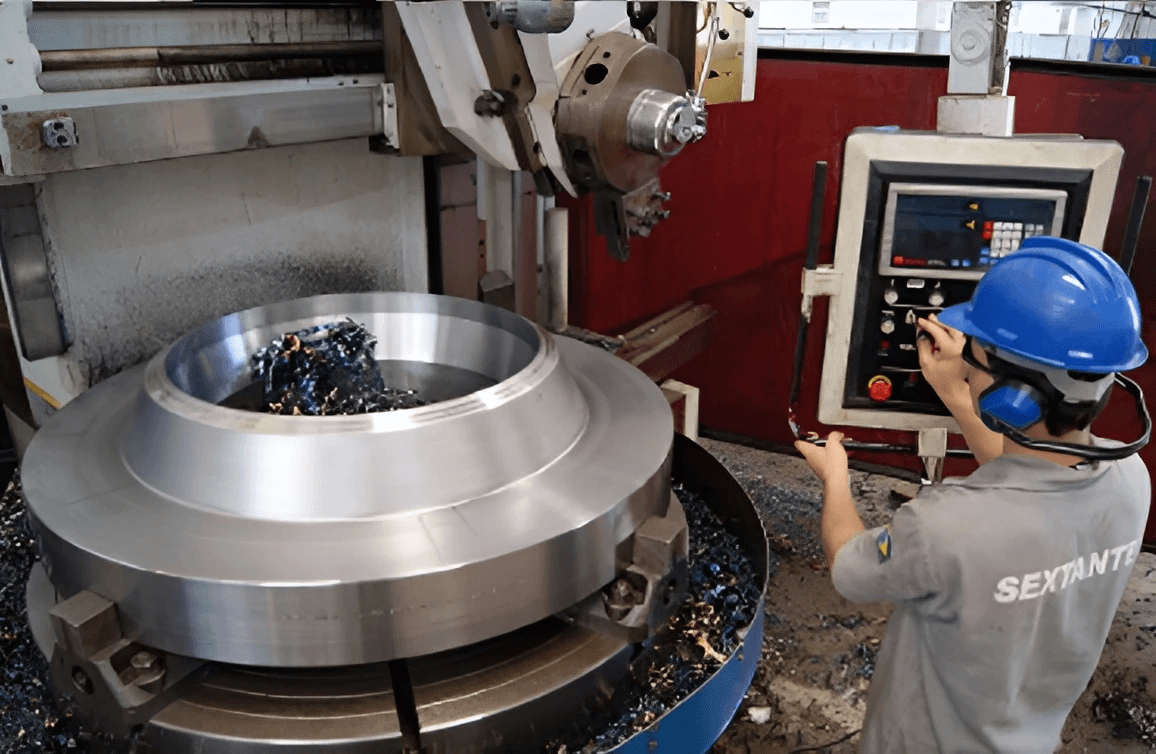 Operador utilizando furadeira radial industrial em processo de perfuração de peça metálica de grande porte.